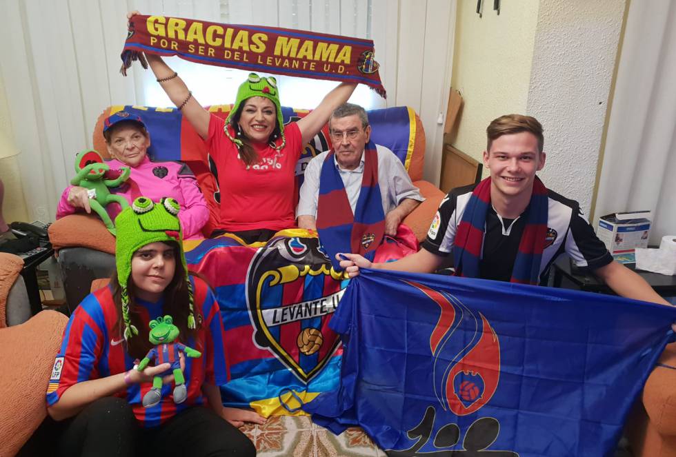 La abuela Dolores Payá es la responsable de que que la familia de Empar Piera sienta pasión por el Levante Unión Deportiva. Esta valenciana del Cabanyal acompañaba al equipo allá donde fuera. “Ella tenía el pase en primera fila, justo frente a la tribuna. Al llegar, desplegaba una toalla con el escudo del club”, relata su nieta Empar, de 53 años, en conversación telefónica. Doloretes, como la conocían sus vecinos, era una mujer inquieta que disfrutaba viendo todo tipo de deportes. “Estaba adelantada a su tiempo”, apunta Empar. Aunque ya no está entre los suyos —falleció a los 84 años en 1989—, dejó su estampa profundamente marcada en el corazón de Empar. Ahora, esta funcionaria del Ayuntamiento de Valencia es la columna vertebral de la familia granota, que siempre, desde los tiempos mozos de Doloretes, ha estado muy ligada al equipo. “De joven, mi padre jugó en uno de los equipos que nutrían a la cantera del Levante”, recuerda.   Desde que tiene uso de razón, Empar ha vivido intensamente los colores. “Nosotros subimos y bajamos de categoría como un ascensor de El Corte Inglés”, confiesa. Hasta ahora ha acudido al estadio Ciutat de València con sus padres y su hija Marina. Todos son socios y abonados, aunque su madre ha dejado de ir y su padre acude con menos frecuencia debido a sus problemas de salud. Cuando van, se sientan en la grada de Orriols, desde la que su padre tiene mejores vistas del campo. Marina promete ser tan aficionada como su bisabuela. La joven, de 21 años, goza de todo lo que tiene ver con el club granota y no se pierde un partido junto a su madre cuando el Levante juega en casa. Su bisabuela, Doloretes, estaría orgullosa.   En la foto: Empar Piera (centro) junto a sus padres, Carlos Piera y Amparo Beltrán, su hija Marina y su sobrino, César Nebot, en el salón de su casa apoyando al Levante Unión Deportiva.